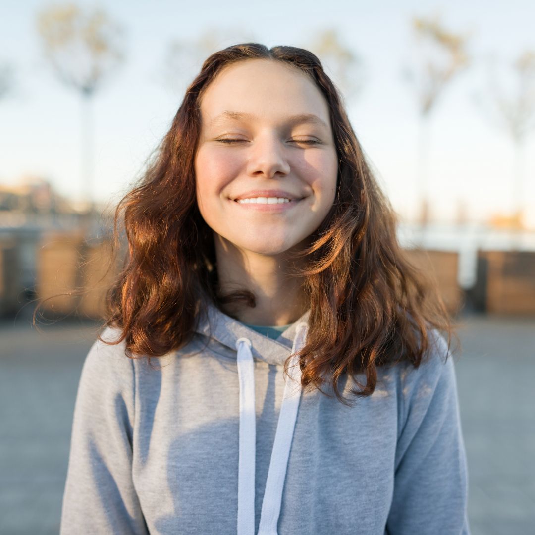 Joyful Moment Outdoors A young woman with wavy brown hair smiles warmly with her eyes closed, standing in a serene outdoor setting.