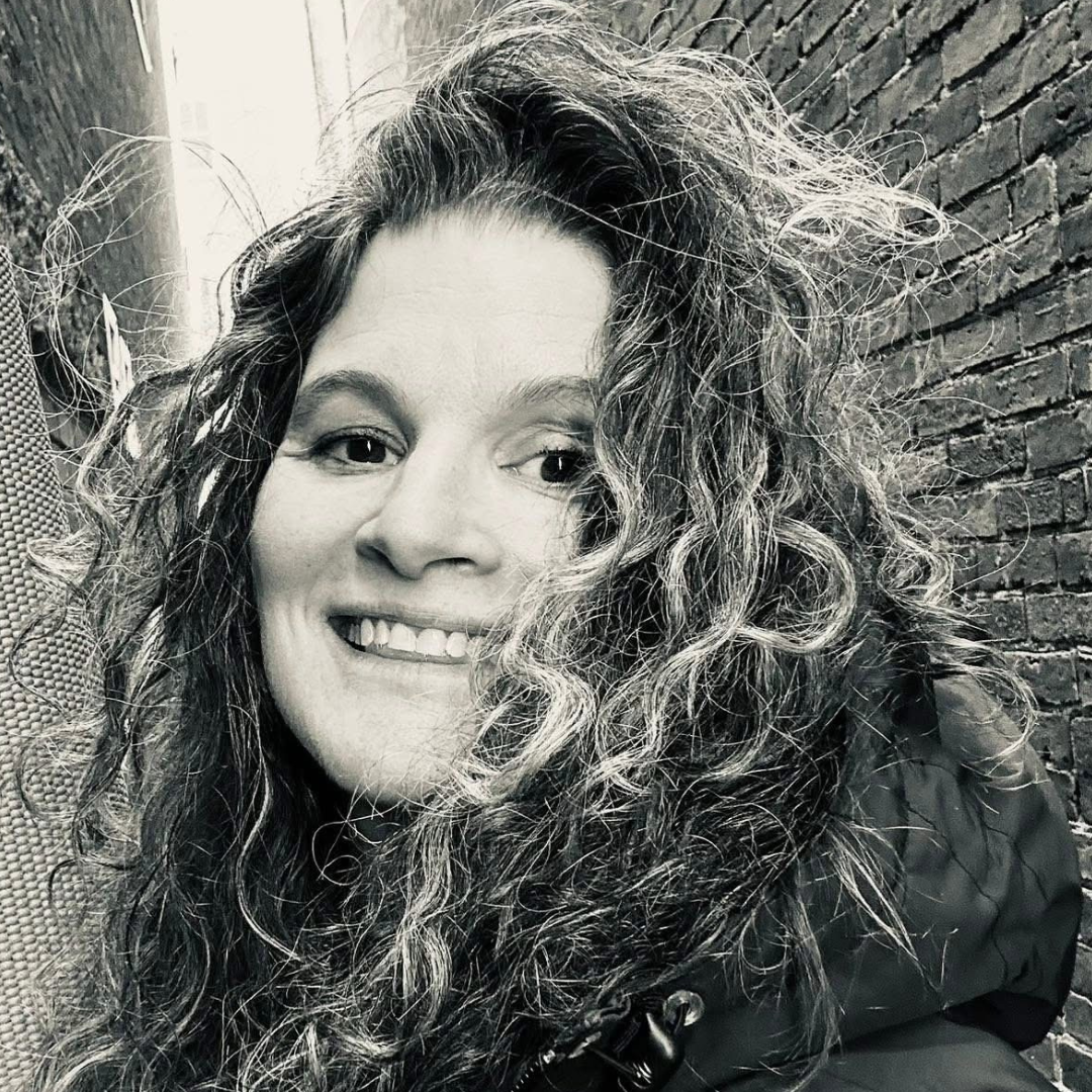A woman with curly hair is smiling while posing in a narrow alleyway, surrounded by brick walls, captured in black and white.